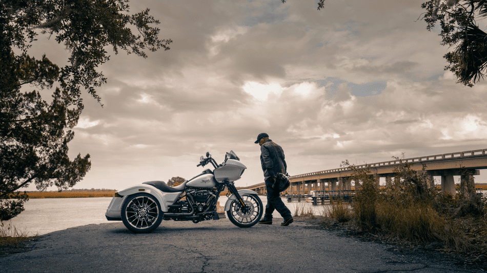 Rider walks toward the 2025 Harley-Davidson® Road Glide® 3 near a lakeside bridge in Deer Park, TX—showcasing its custom touring style.